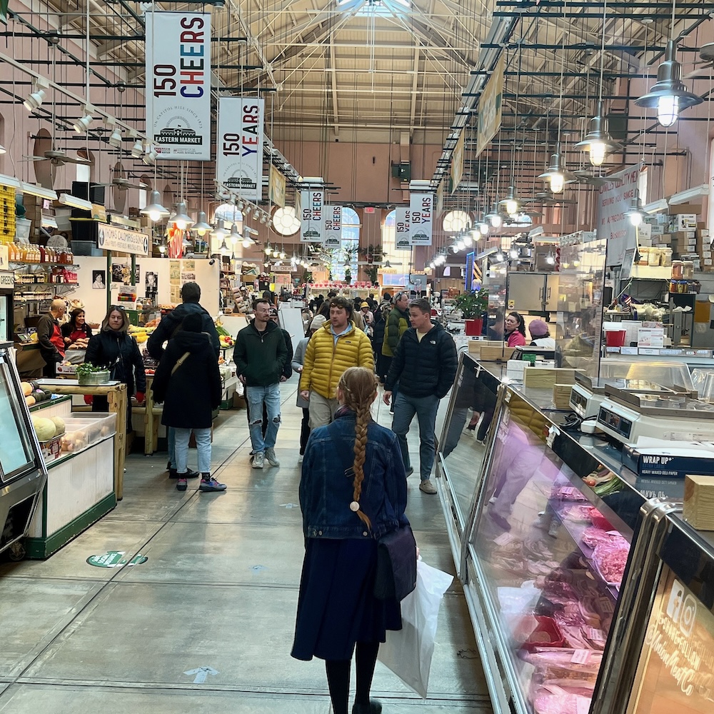 a teen walks past a butcher counter and fresh produce as she passed through the main market building of eastern market.