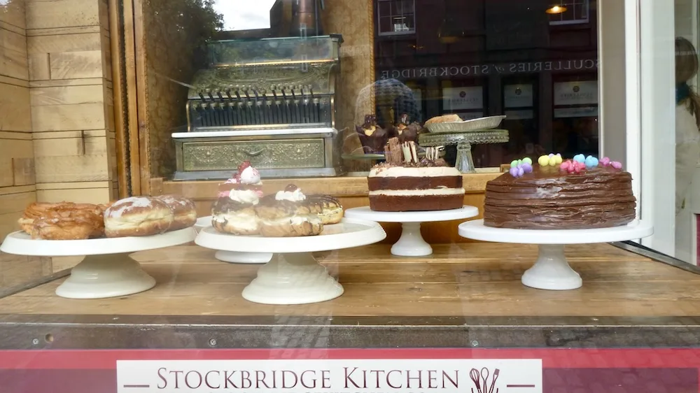 a window full of cakes, cream puffs and an old-fashioned cash register on the stockbridge high street in edinburgh.